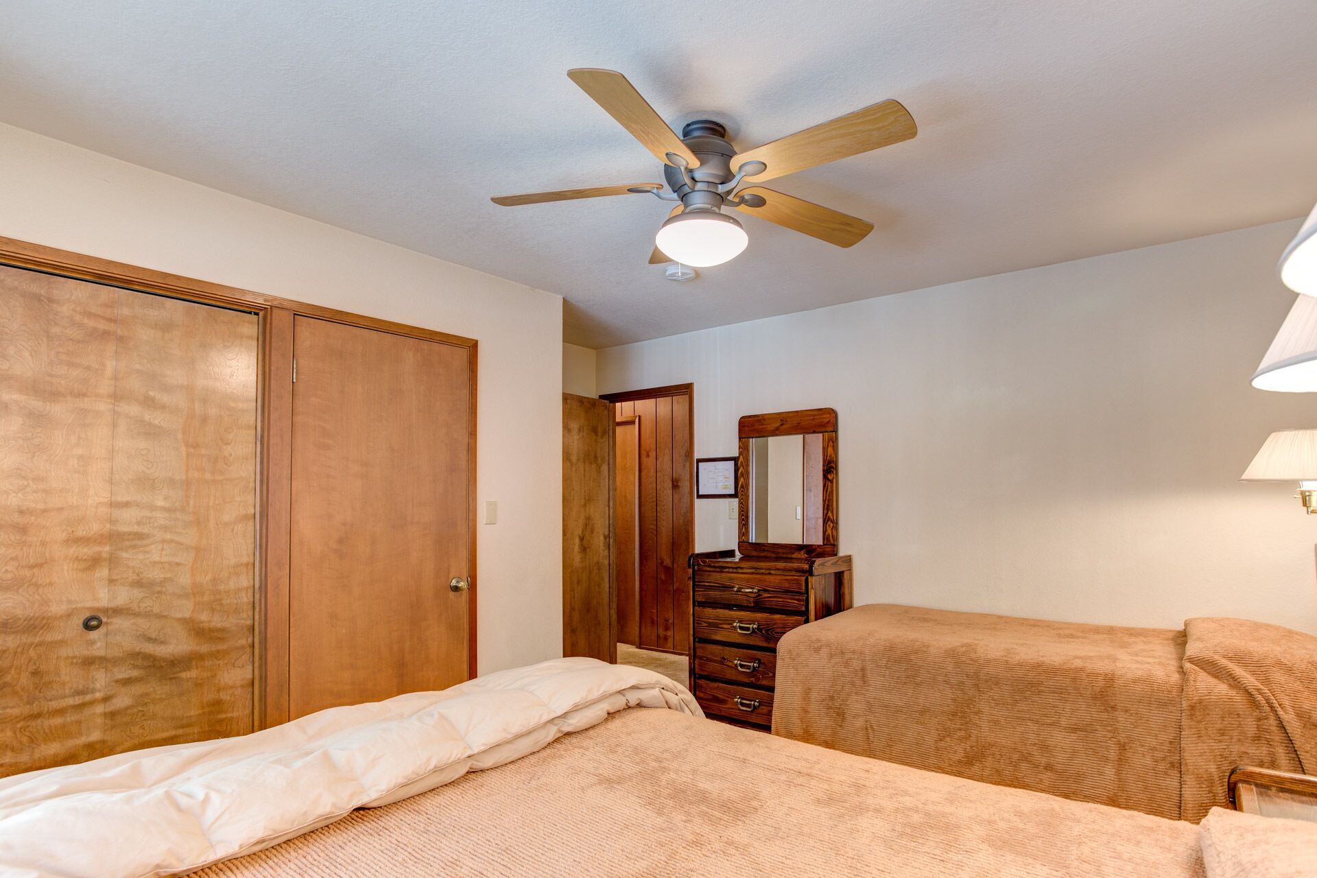 A bedroom with a wooden wardrobe, a dresser, a bed with a beige blanket, and a ceiling fan.