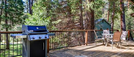 Una parrilla para barbacoa en una terraza de madera con vistas a un bosque y una casa al fondo.
