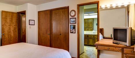 A bedroom with a bed, wooden wardrobe, a vanity with a mirror and lights, and a television.