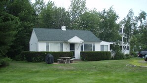 A single-story house with a green roof, a covered patio table, and a hedge in front. - Located In The Heart Of Meredith Bay (Meredith)