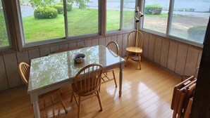 A sunlit room with a glass table and wooden chairs, large windows offering a view of a grassy area and a body of water. - Located In The Heart Of Meredith Bay (Meredith)