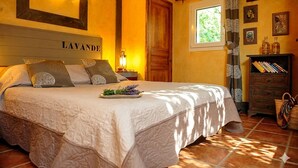Une chambre à coucher comprenant un lit, une table de chevet, une commode, une fenêtre et une étagère fixée au mur, sur laquelle sont posés des livres et des bouteilles.