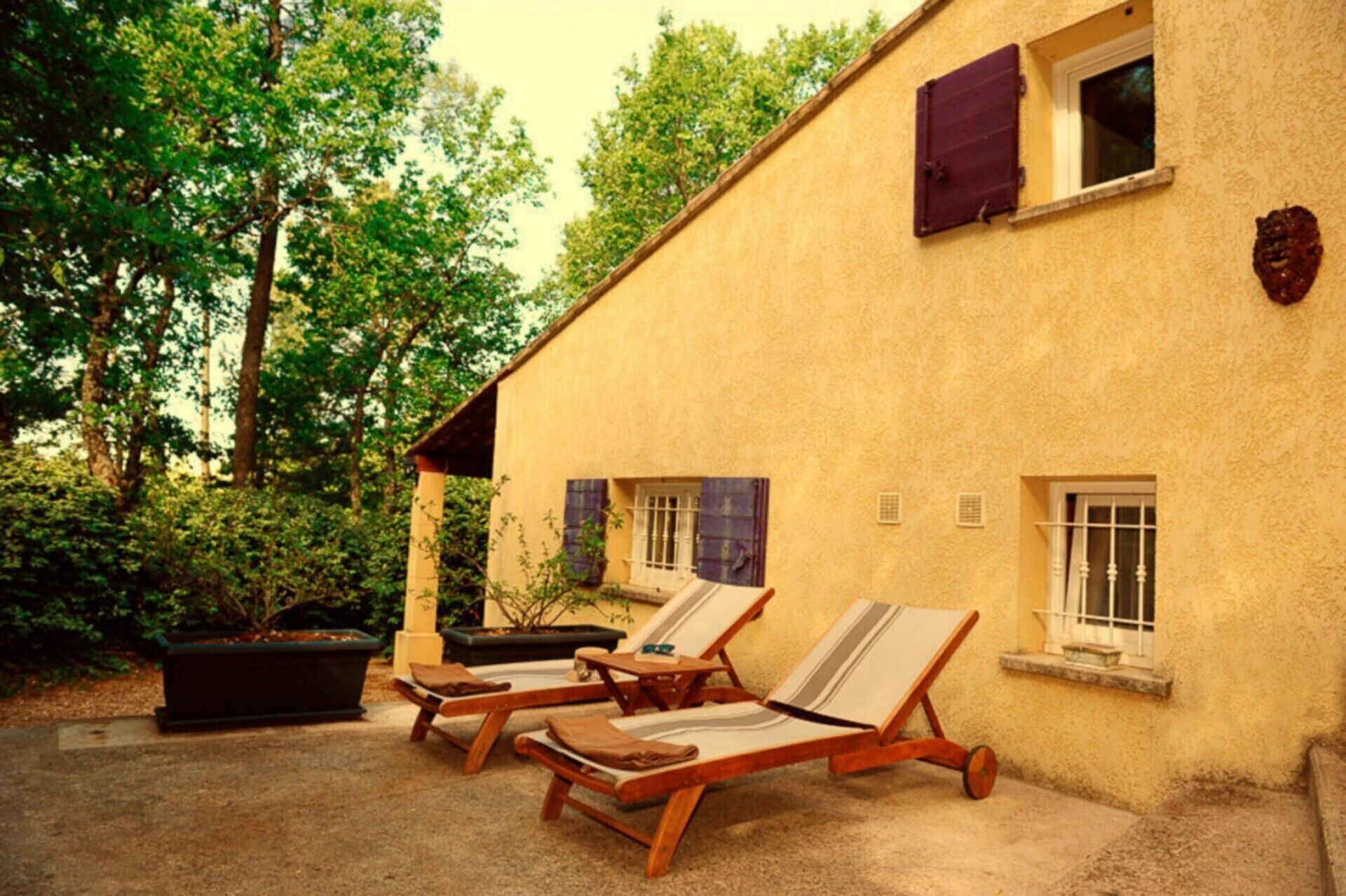 Un bâtiment à la façade jaune, doté de deux fenêtres avec volets et d’un petit balcon. On y trouve deux fauteuils de jardin en bois, équipés de coussins.