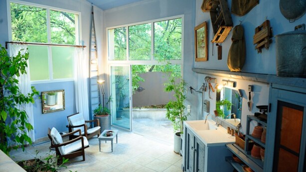 Une pièce spacieuse avec de grandes fenêtres, une porte-fenêtre coulissante donnant sur un balcon, ainsi qu’une variété de plantes et de meubles.
