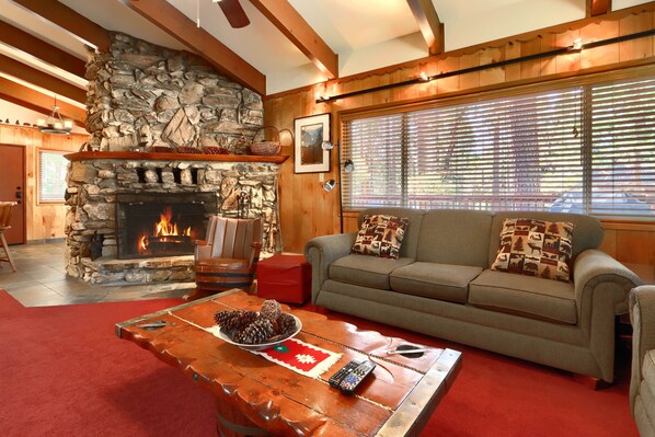 A cozy living room with a stone fireplace, a sofa with patterned cushions, and a wooden coffee table with a remote control.