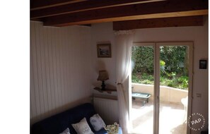 A cozy room with a wooden ceiling, a blue sofa with decorative pillows, a table lamp, and a view of a garden through a sliding glass door.
