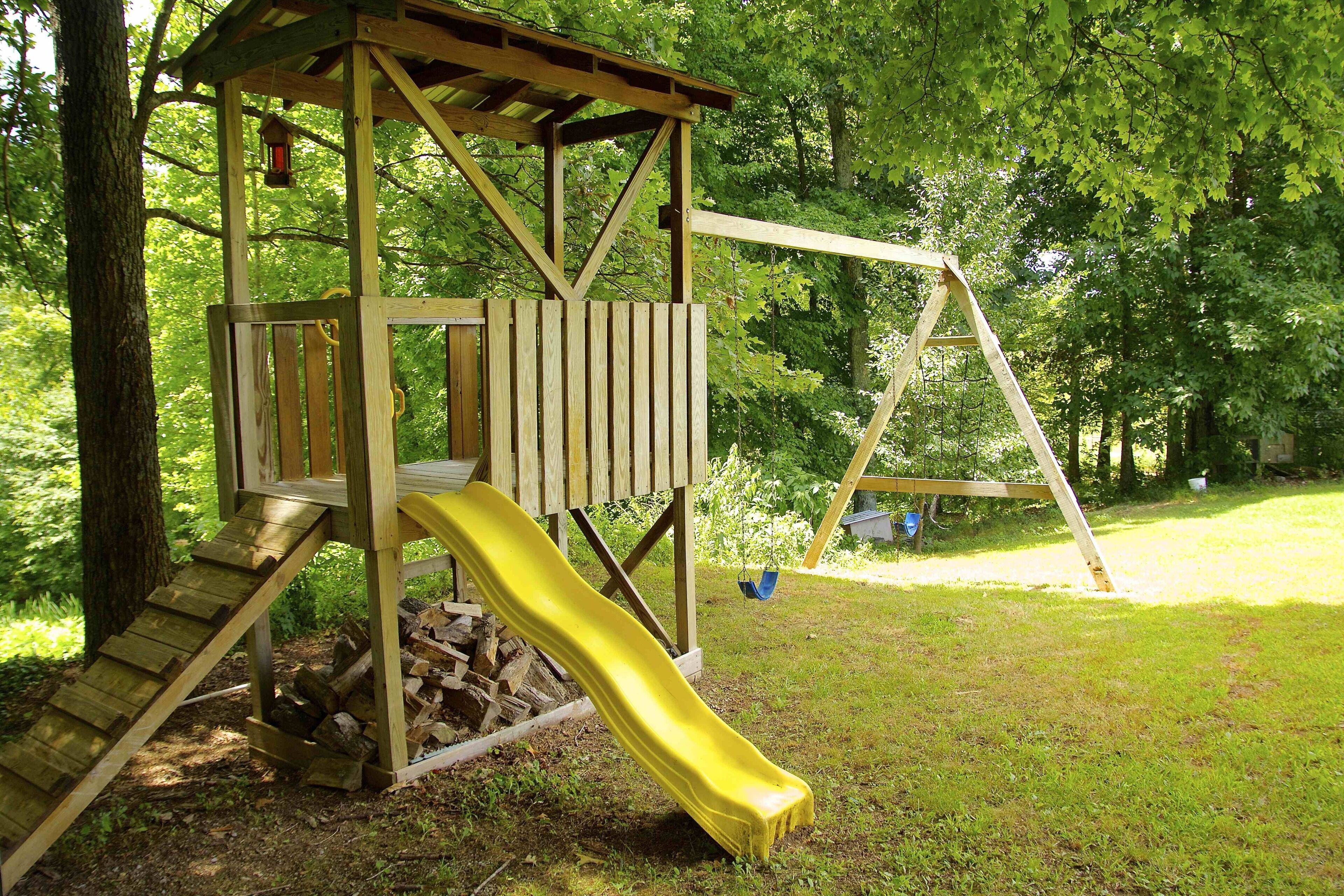 A wooden play structure with a slide and a swing set in a grassy area surrounded by trees.