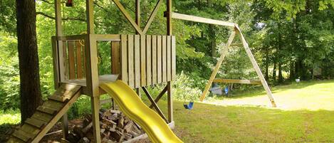 A wooden play structure with a slide and a swing set in a grassy area surrounded by trees.