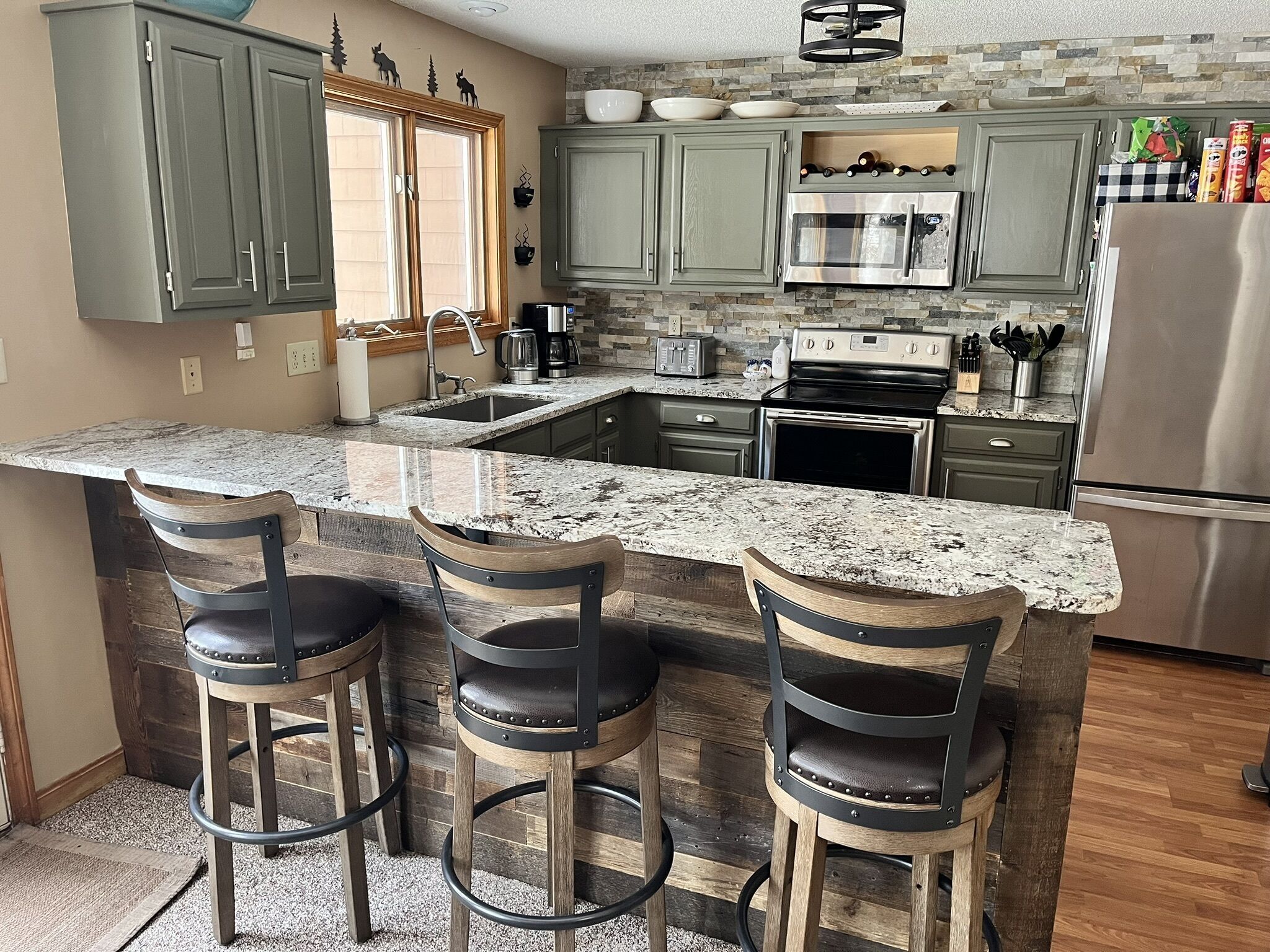A kitchen with a granite countertop, green cabinets, a stainless steel refrigerator, and three bar stools.
