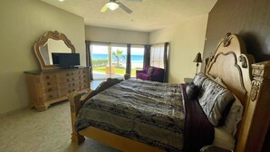 Een slaapkamer met een groot bed, een kapstok, een televisie en uitzicht op de oceaan door grote ramen.