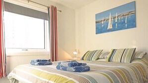 A hotel room with a large bed, striped bedding, a painting of sailboats on the wall, and a window with curtains.