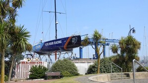 A sailboat with '11th Hour Racing' written on it, docked near a waterfront with palm trees and a bridge in the background.
