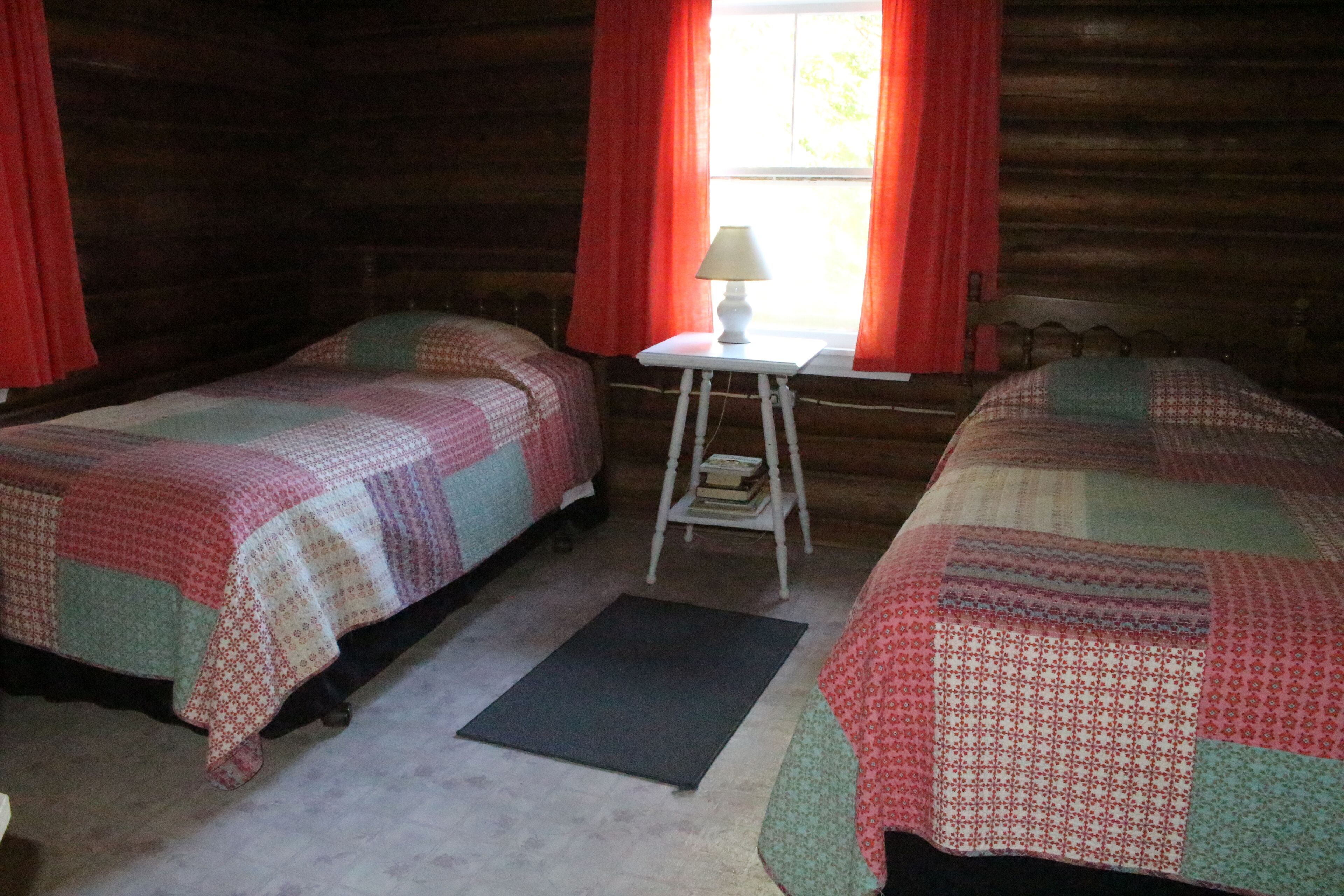 A cozy cabin room with two beds, a small table with a lamp, and a window with red curtains.
