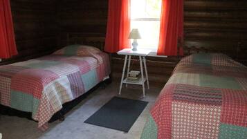A cozy cabin room with two beds, a small table with a lamp, and a window with red curtains.