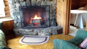 Un camino in pietra con un fuoco acceso, una panca di legno e un letto con coperta bianca.