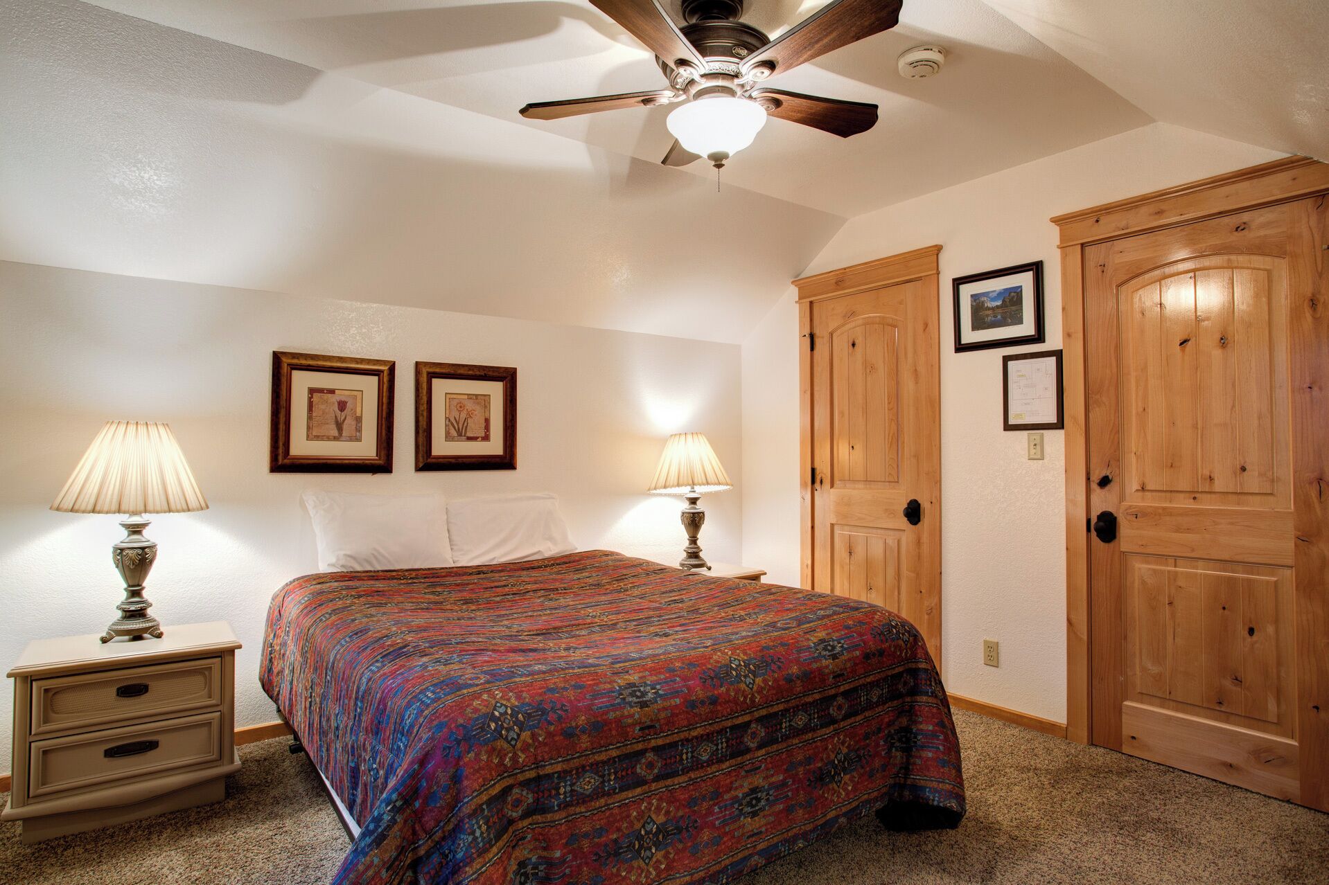 A bedroom with a bed, two bedside lamps, a ceiling fan, and wooden doors.