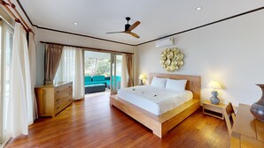 Ein Schlafzimmer mit einem hölzernen Bett, Deckenventilator, Klimaanlage, Nachttisch mit Lampe und Blick auf einen Balkon mit einer Sitzecke.
