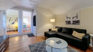 Un salon avec un canapé en cuir noir, une table basse en verre et une lampe. Une porte donne accès à une chambre à coucher qui contient un lit et un bureau.