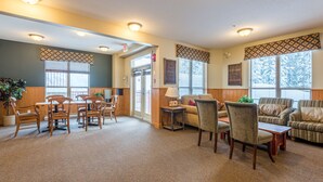 A spacious living area with a carpeted floor, wooden furniture, and large windows with patterned valances.