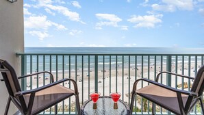 A balcony with two chairs and a small table, overlooking a beach with people and umbrellas. - Caribbean Paradise-Romantic Oceanfront Canopy King Suite with Breathtaking Views (Myrtle Beach)