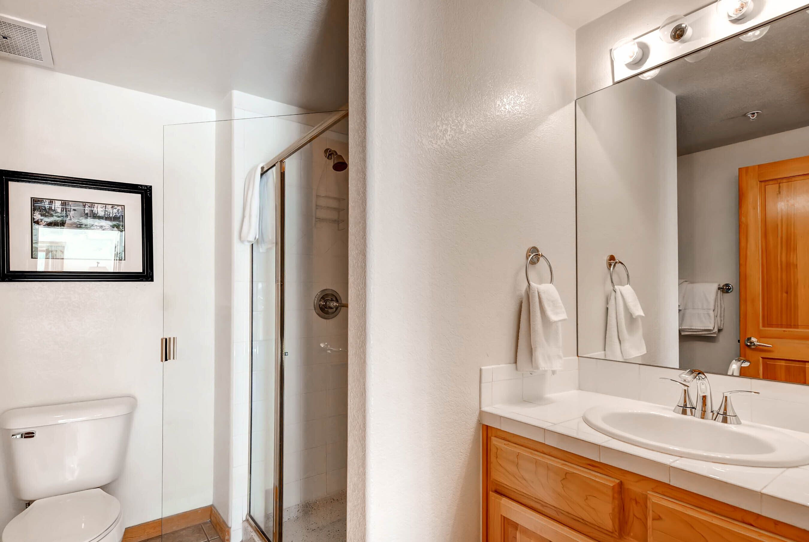 A bathroom with a glass shower enclosure, a white toilet, a wooden vanity with a sink, and a large mirror.