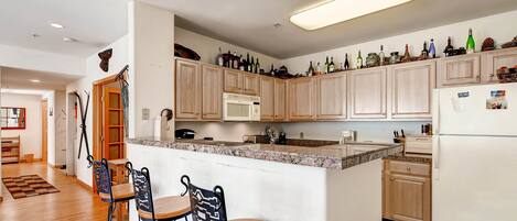 A kitchen with wooden cabinets, a granite countertop, and a refrigerator. There are two bar stools and a microwave.