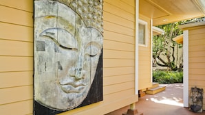 A building exterior with a large Buddha statue on the wall, a covered porch, and a small garden area. - Luxury, AC, Adjacent to Acres of State Oceanfront Land (Pahoa)
