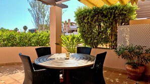 Une table ronde surmontée d’un centre de table en verre, entourée de chaises noires, est placée sur une terrasse carrelée, au-dessus de laquelle se trouve un auvent jaune.