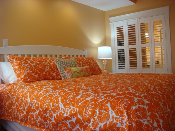 Una cama con una colcha naranja y blanca estampada, un cabecero blanco y una lámpara de noche.