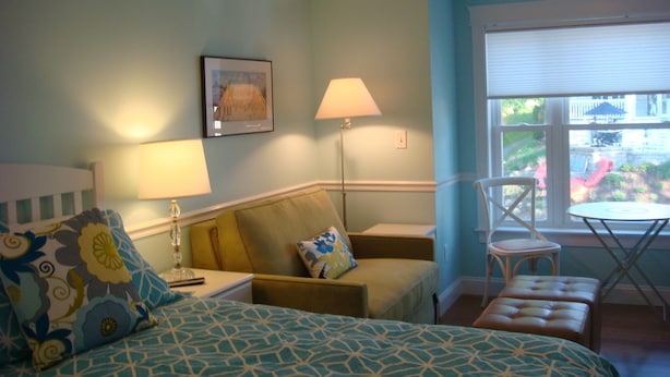 Une chambre à coucher comprenant un lit, deux tables de chevet, une chaise, une petite table et une fenêtre avec des stores.