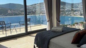 A bedroom with a large bed, a view of a coastal town, and a balcony with chairs.