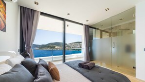 A modern bedroom with a large bed, a glass shower, and a view of the sea.
