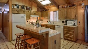 A kitchen with wooden cabinets, a central island with stools, a refrigerator, a microwave, and a window with curtains. - (69) Bordentown West at The Redwoods In Yosemite (Yosemite National Park)