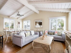 Un salon doté d’un ventilateur de plafond, un coin repas avec une table et des chaises, et une grande fenêtre avec des stores.