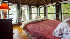 Une chambre avec un plafond en bois, un grand lit recouvert d’une courtepointe rouge, une table de chevet avec une lampe et de grandes fenêtres aux cadres verts.