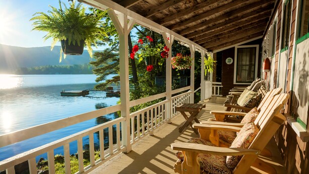Une terrasse en bois dotée d’une rambarde, qui offre une vue sur un lac, avec un bateau sur l’eau et des plantes en pot suspendues au plafond.