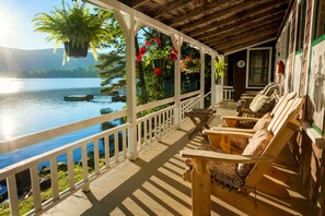 Une terrasse en bois dotée d’une rambarde, qui offre une vue sur un lac, avec un bateau sur l’eau et des plantes en pot suspendues au plafond.