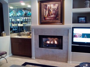 A living room with a fireplace, a television, and a bar area with glassware. - VENEZIA - Heated Pool/Spa - Large Modern Single Story - 3BR, 3 BA 3100 Sq. Ft (Palm Desert)
