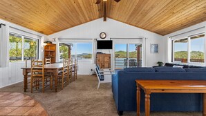 Un vaste espace de vie doté d’un plafond en bois, d’un canapé bleu, d’une table à manger avec des chaises et de grandes fenêtres offrant une vue sur l’extérieur.