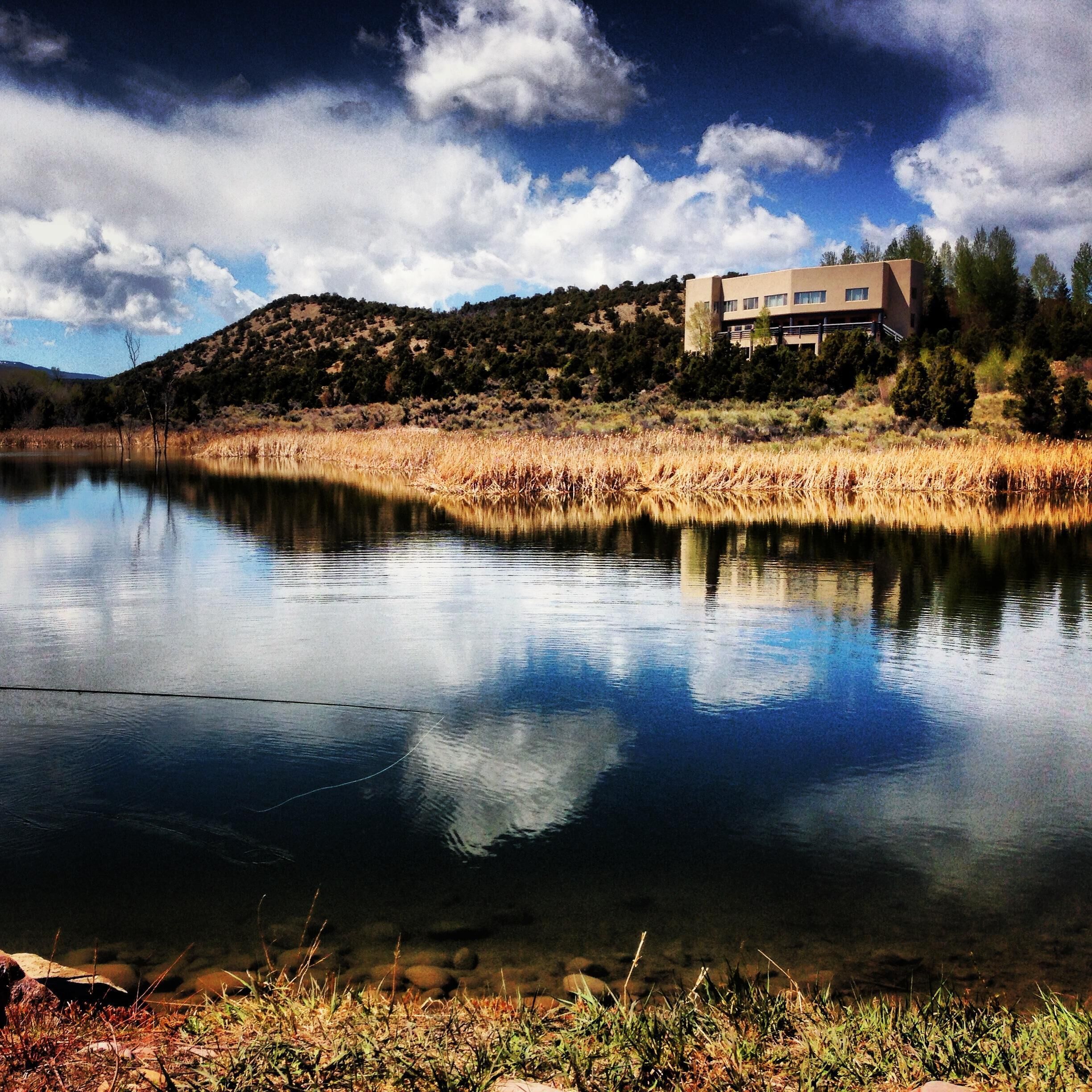 A modern building with a flat roof, surrounded by a natural landscape with a body of water reflecting the sky and clouds.