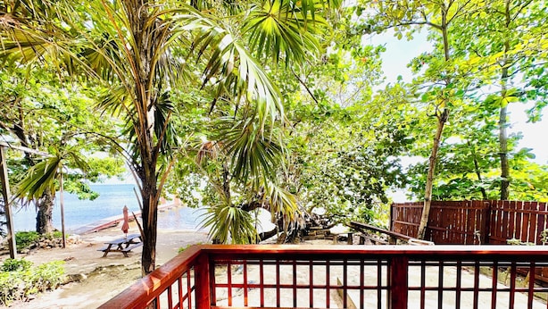 A wooden deck with a railing overlooking a beach and palm trees. - Stay & Dive! Ocean front, Cozy Casita steps to the waters edge!  (Roatan, Bay Islands)