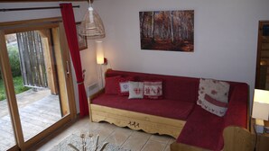 A cozy room with a wooden ceiling, a red sofa with patterned cushions, a painting of a forest, and a large window with a view of greenery. - Light airy Chalet - Mountain And Waterfall Views. Excellent access all Amenities (Samoëns)