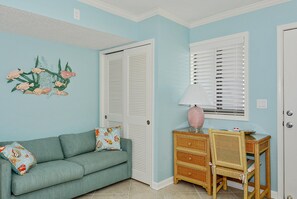 Une pièce avec un canapé de couleur turquoise, une petite table d’appoint en osier surmontée d’une lampe, un mur orné d’un tableau floral et une porte dotée de volets à persiennes.