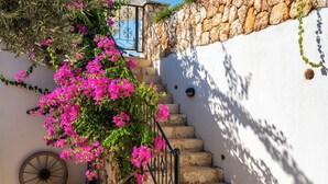 Una escalera de piedra con barandilla de hierro forjado que sube hacia una pared blanca, junto a otra pared de piedra y una puerta azul.