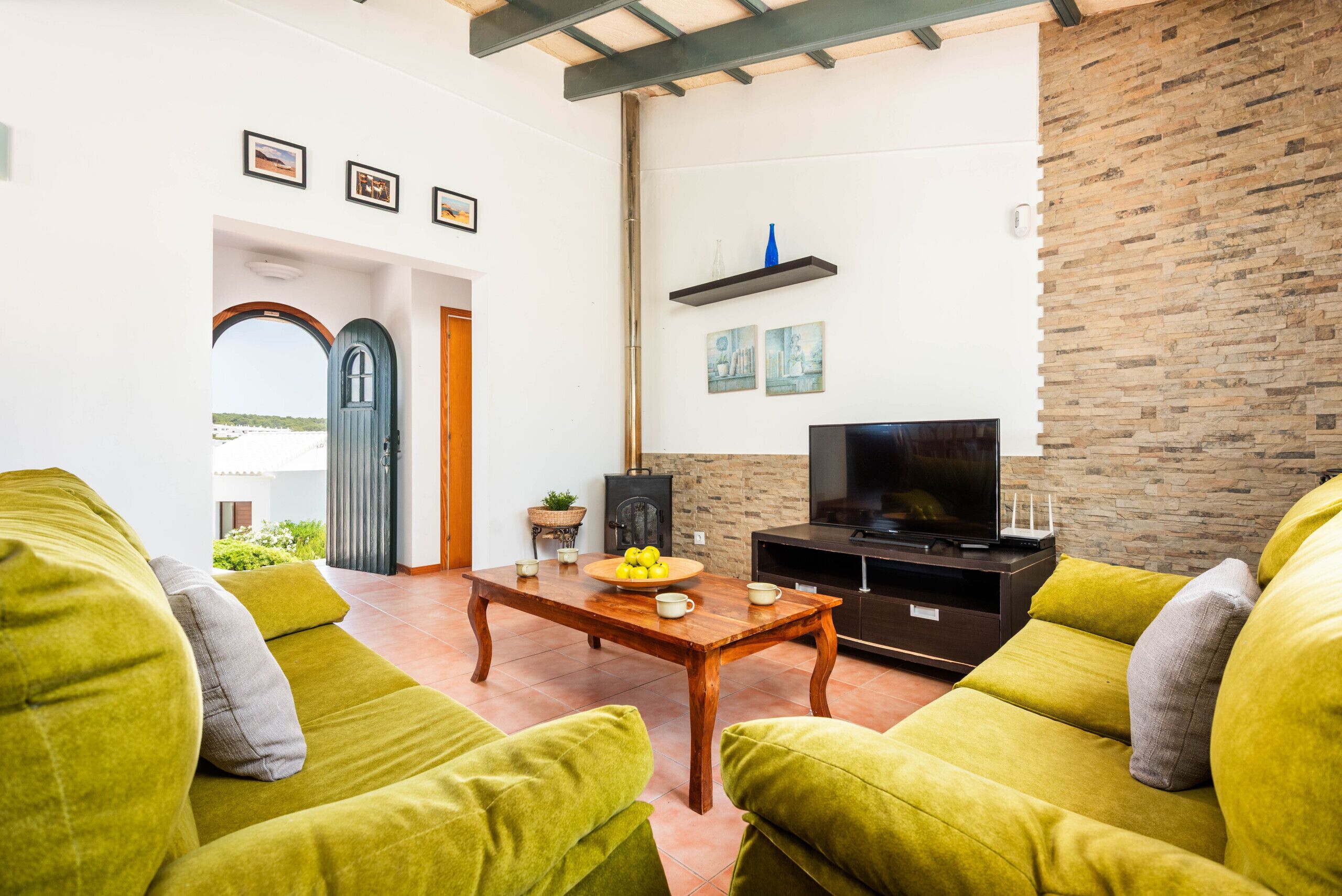 Una sala de estar con una mesa de centro de madera, sofás verdes, un televisor y una pared de piedra.