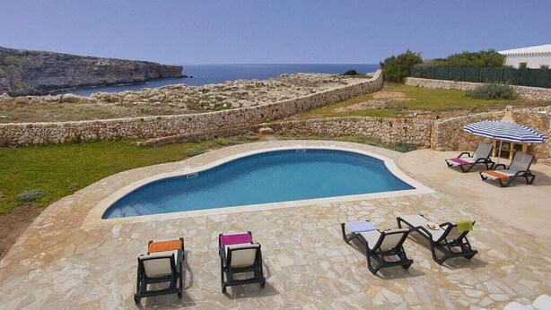 Une piscine aux bords recouverts de pierres, entourée de chaises longues et d’un mur en pierre.