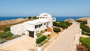 Une maison blanche située sur la côte, avec une allée incurvée, un escalier menant à l’entrée et une vue sur la mer.