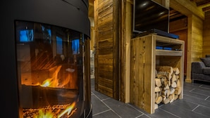 Een houtkachel met zichtbare vlammen, een houten deur en een stapel haardhout in een kamer met houten wanden en een betegelde vloer.