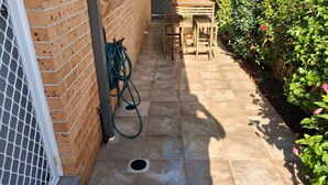 A patio area with a brick wall, a metal gate, a wooden table and chairs, and a garden with green foliage and a red flower. - Beach Haven Villa - close to beach and breakwall (NORTH HAVEN)