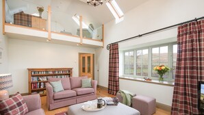 A cozy living room with a sofa, ottoman, and a coffee table with a tray. There is a bookshelf and a window with curtains. - TIME - Charming Countryside Barn Conversion with Hot Tub & Scenic Views (Galashiels)